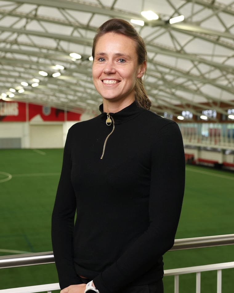 Cara Lea Moseley, founder of Virtuoso Psychology, standing pitchside at an indoor training facility, smiling to camera.